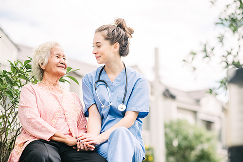 Young nurse in light blue scrubs sitting outside with elderly asian woman holding her hands